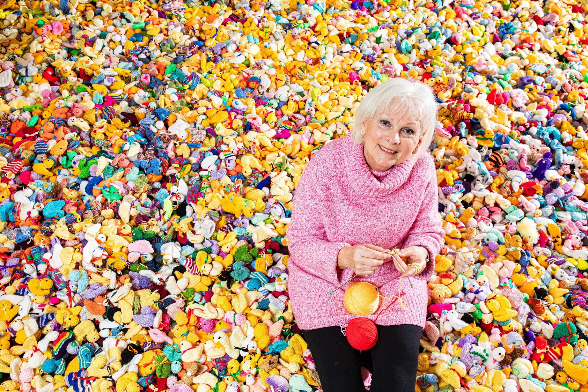 Woman wearing a pink jumper knitting sat on the floor surrounded by thousands of colourful knitted chicks