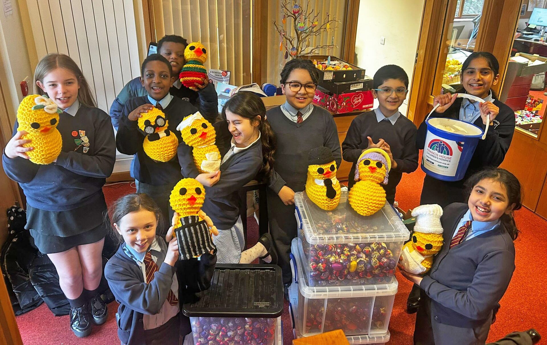 Children in school uniform pose with giant knitted chicks amongst boxes full of Creme Eggs