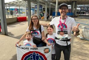 Family of two adults and two children wearing t-shirts and holding a banner with a rainbow logo. The man is wearing a London Marathon running medal. They are smiling and standing outside a train station.