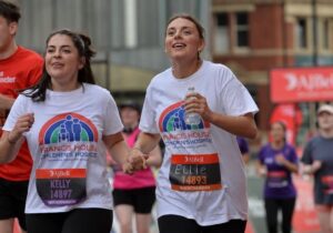 two women wearing white t-shirts with a rainbow logo and the words Francis House children's hospice running in a race in the city streets of Manchester
