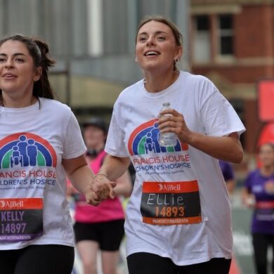two women wearing white t-shirts with a rainbow logo and the words Francis House children's hospice running in a race in the city streets of Manchester