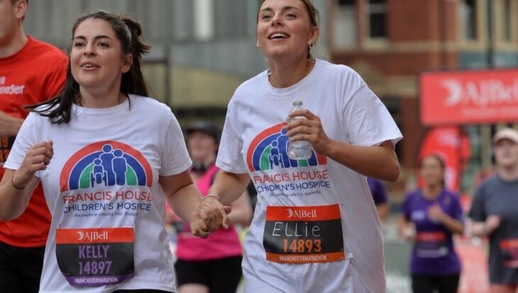 two women wearing white t-shirts with a rainbow logo and the words Francis House children's hospice running in a race in the city streets of Manchester