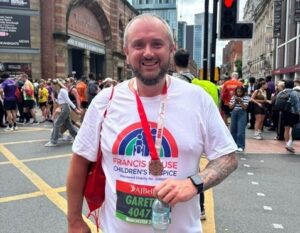 Man with a beard wearing a white t-shirt with a rainbow logo and a running medal around his neck standing on a city street with crowds of people in the background