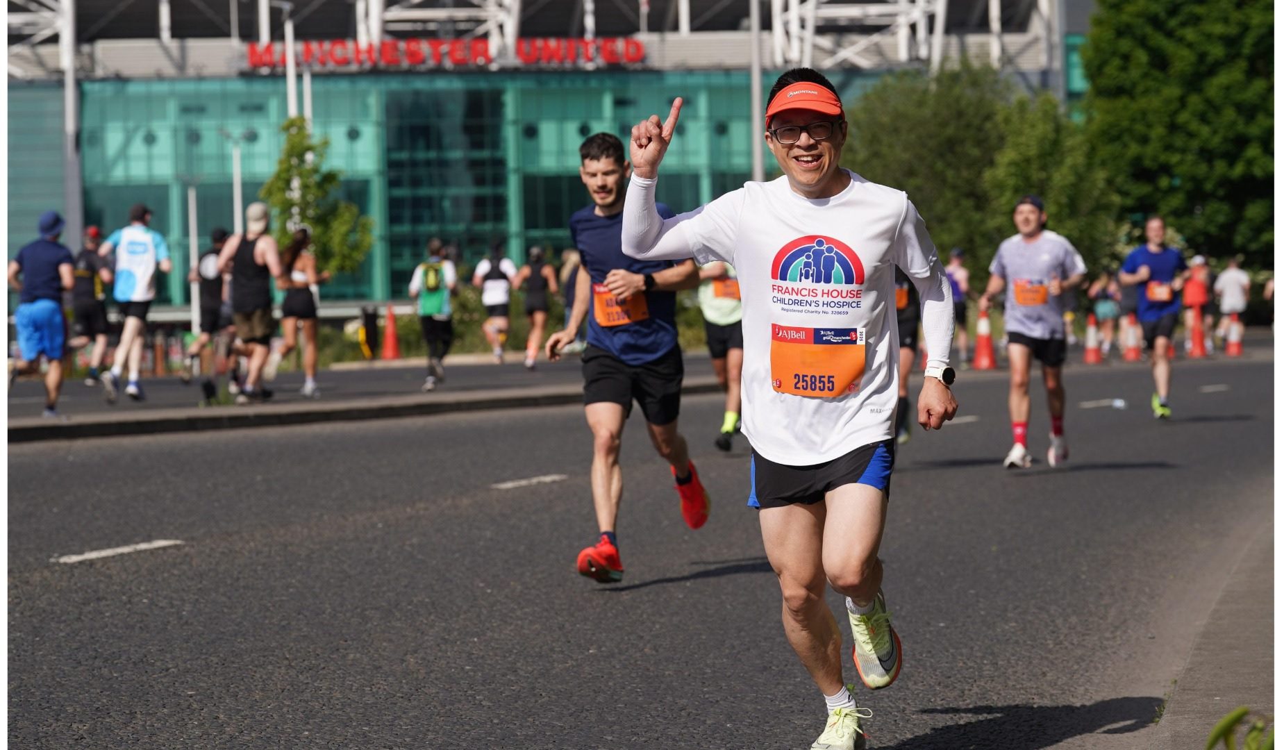 Man wearing running shirt and shorts holds his finger in the air as he runs past Manchester United football ground with other runners in the background.