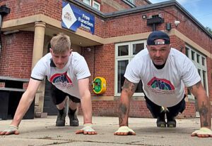 Two men doing push ups outside a pub, they are wearing white t-shirts with a rainbow coloured logo