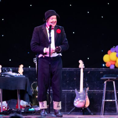 Comedian on a stage wearing a dinner jacket and wheelie boots with L and R drawn on them, surrounded by balloons and props