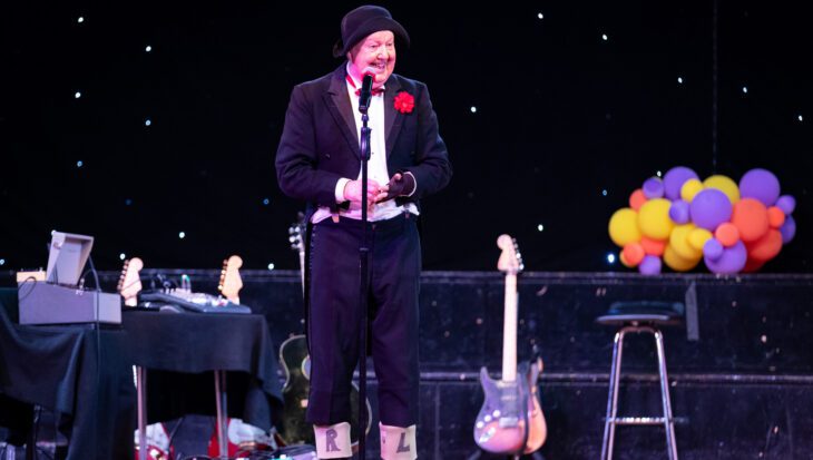 Comedian on a stage wearing a dinner jacket and wheelie boots with L and R drawn on them, surrounded by balloons and props