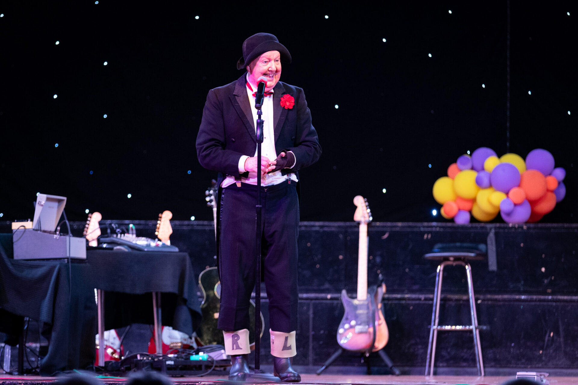 Comedian on a stage wearing a dinner jacket and wheelie boots with L and R drawn on them, surrounded by balloons and props