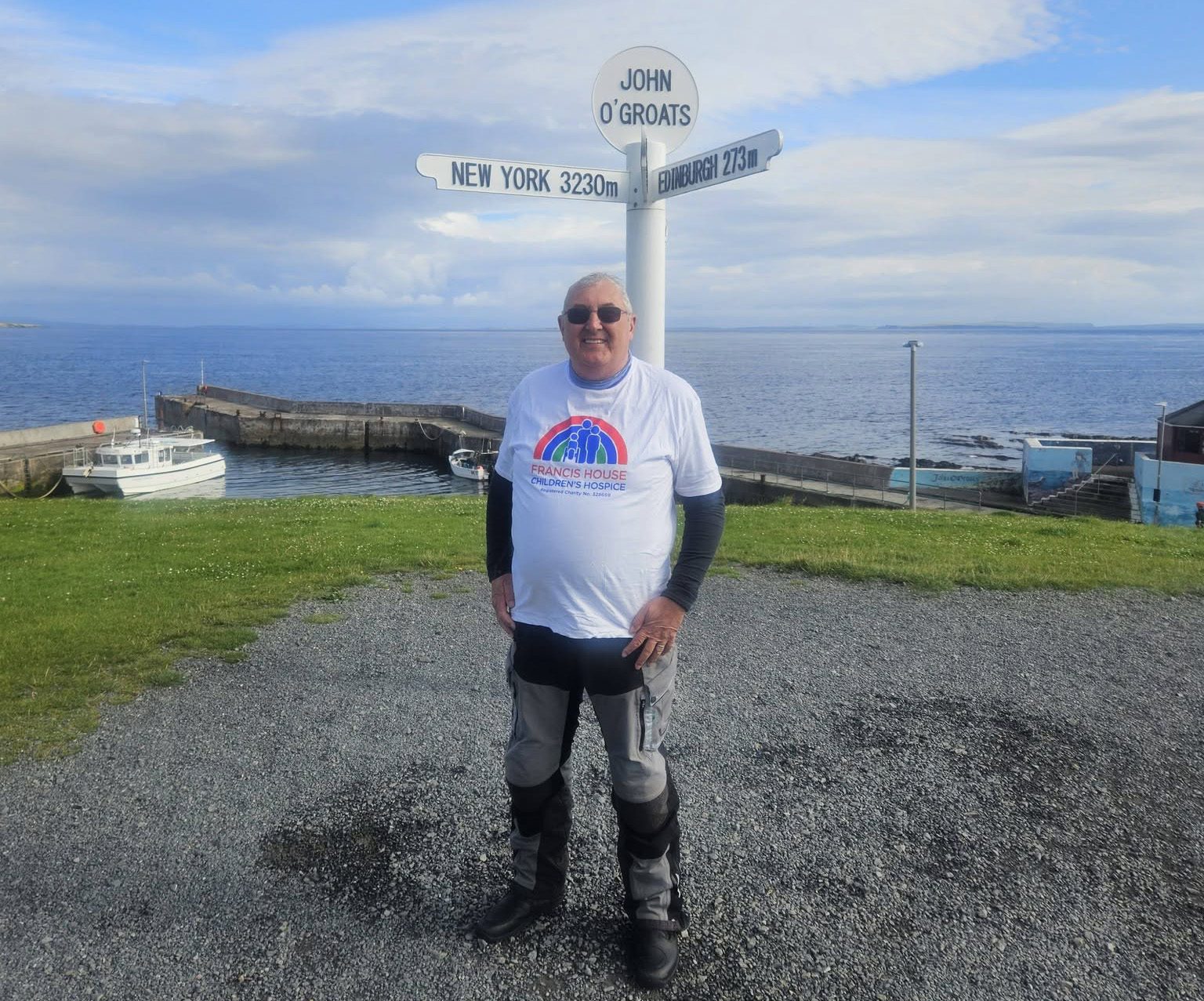 Man wearing a white t-shirt with rainbow logo and biker trousers stood in front of the famous John O'Groats sign in Scotland