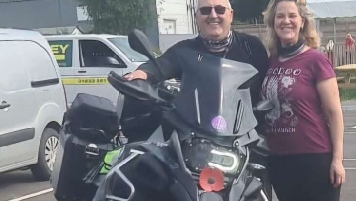 Man and woman stood next to a motorbike in a car park smiling at the camera