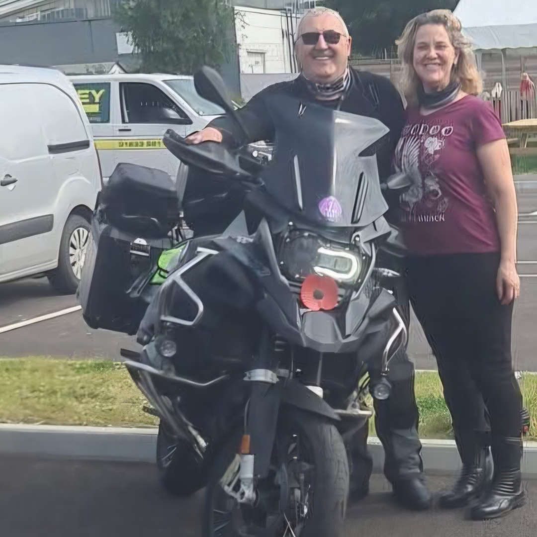 Man and woman stood next to a motorbike in a car park smiling at the camera
