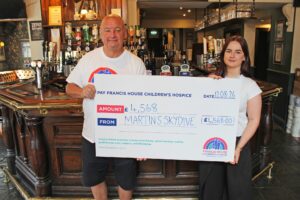 Man and woman stood in front of a bar in a pub holding a large cheque that says Martin's skydive £4,568 to Francis House Children's Hospice