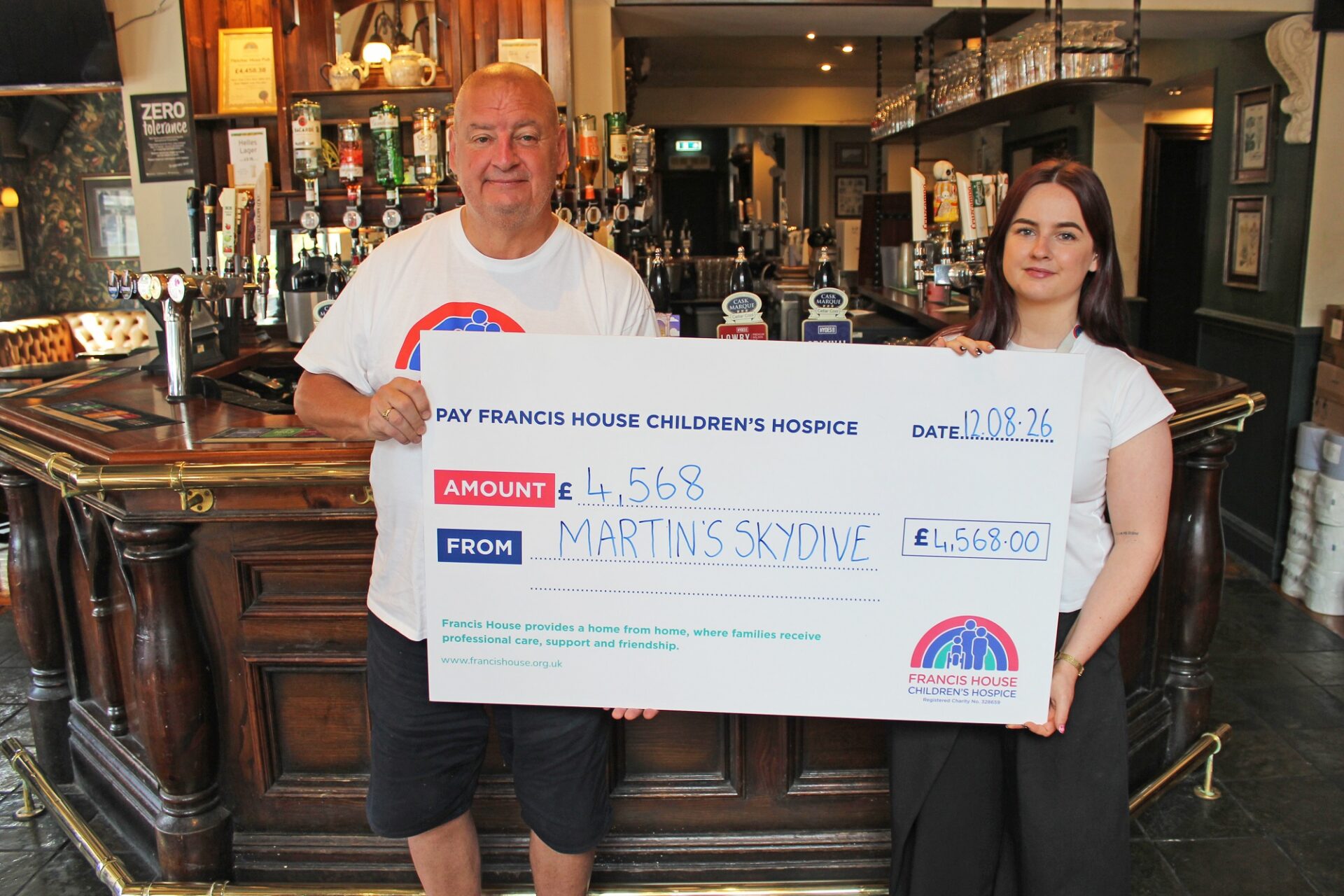 Man and woman stood in front of a bar in a pub holding a large cheque that says Martin's skydive £4,568 to Francis House Children's Hospice