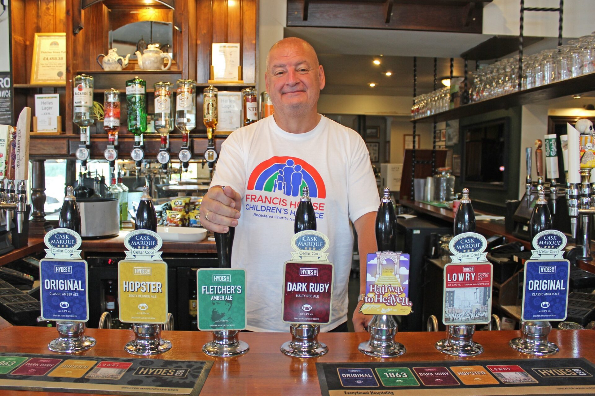 Man wearing a white t-shirt with a rainbow logo is stood behind a bar with his hand on the pump of one of the row of beers