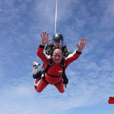 Man wearing a red jumpsuit with his arms raised in the air skydiving in tandem with an instructor wearing a black jumpsuit as they freefall in a blue sky