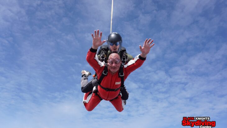 Man wearing a red jumpsuit with his arms raised in the air skydiving in tandem with an instructor wearing a black jumpsuit as they freefall in a blue sky