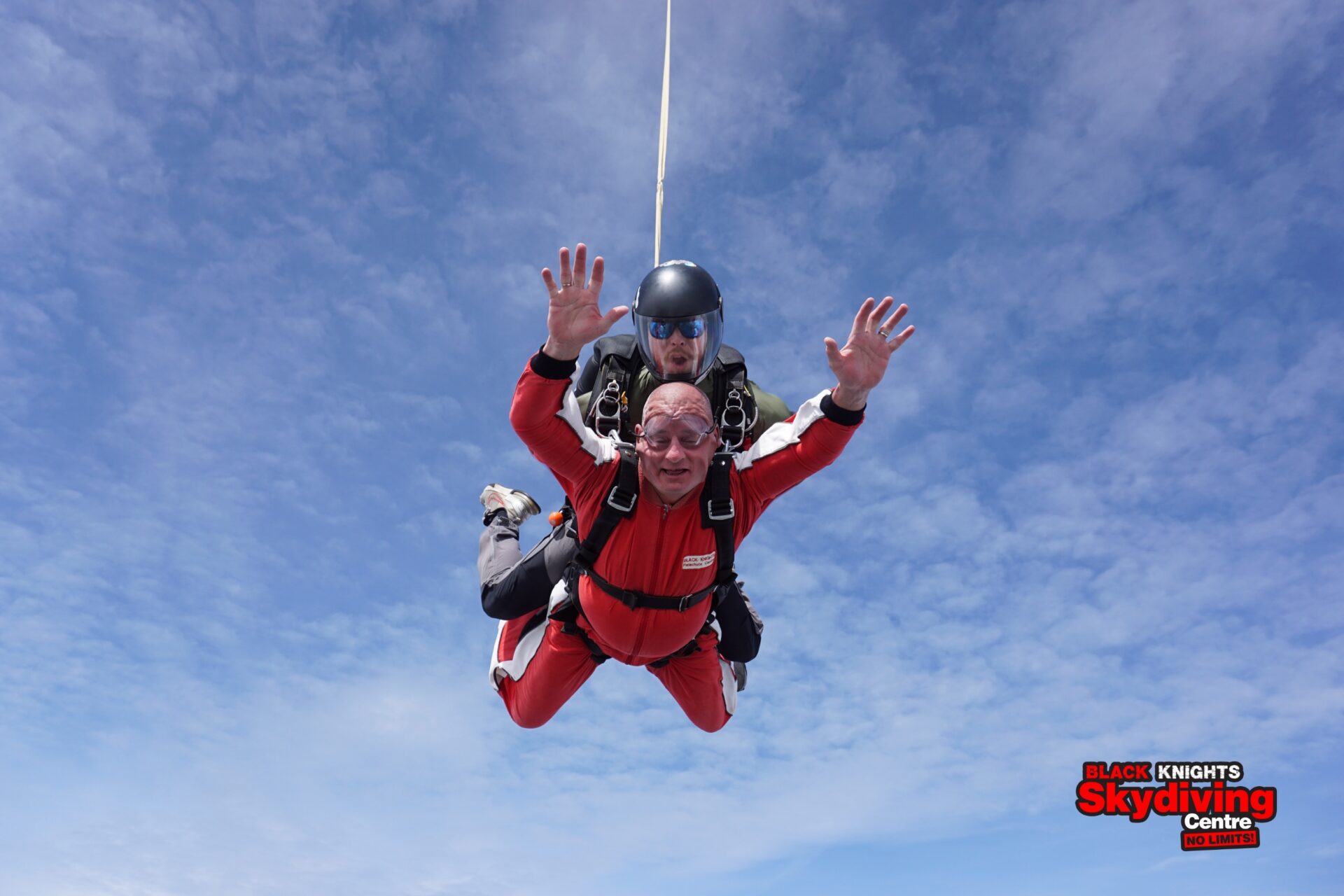 Man wearing a red jumpsuit with his arms raised in the air skydiving in tandem with an instructor wearing a black jumpsuit as they freefall in a blue sky