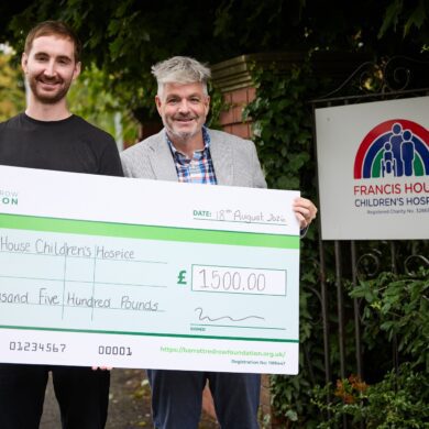Two men stood holding a large cheque donation to Francis House children's hospice stood by an entrance gate