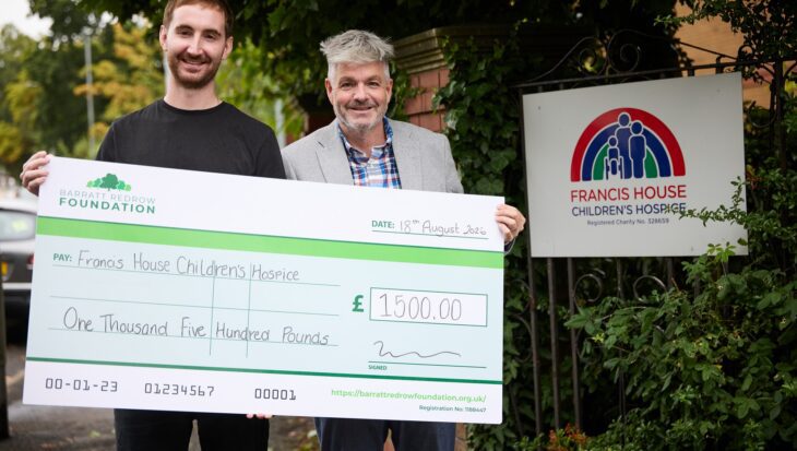 Two men stood holding a large cheque donation to Francis House children's hospice stood by an entrance gate