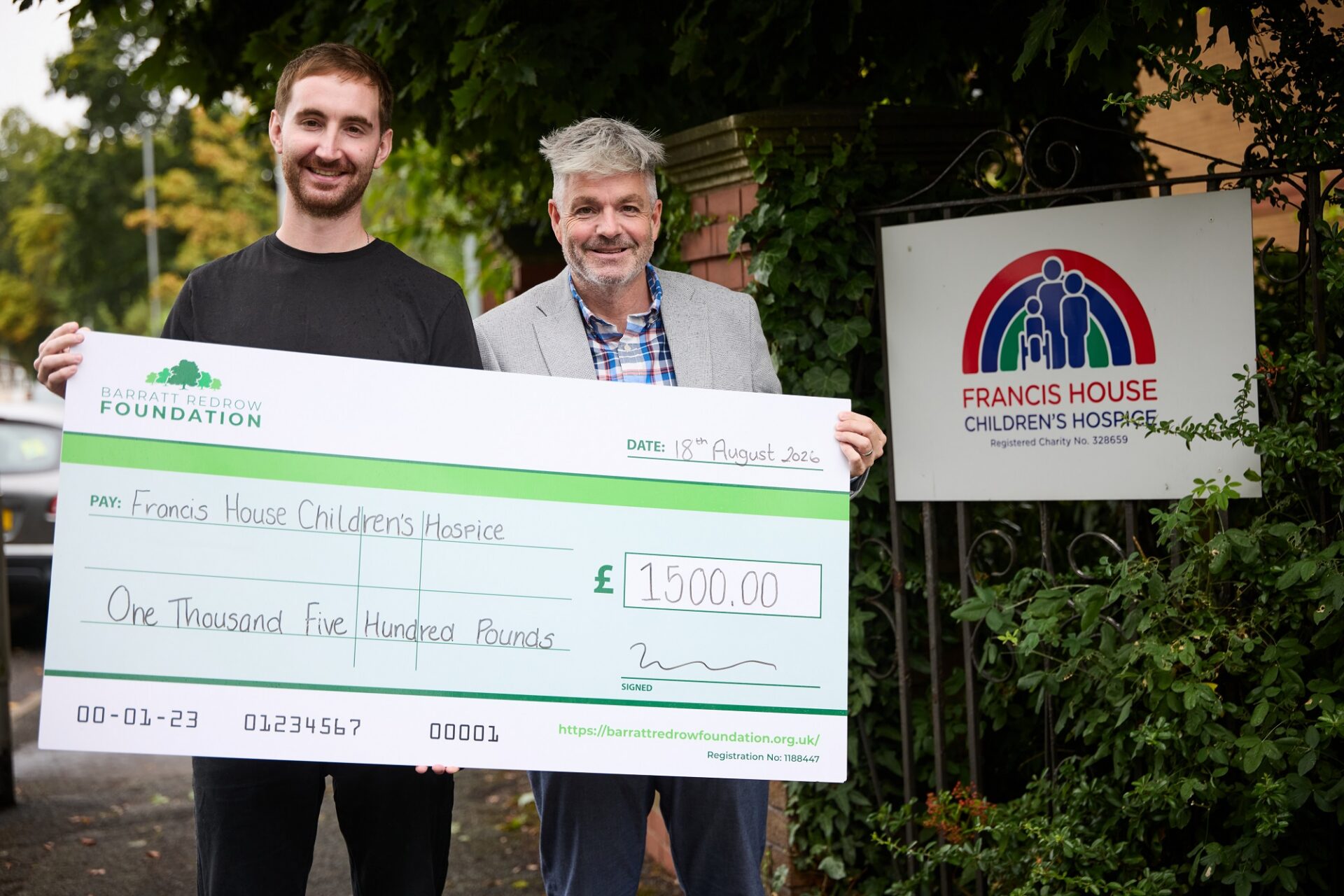 Two men stood holding a large cheque donation to Francis House children's hospice stood by an entrance gate