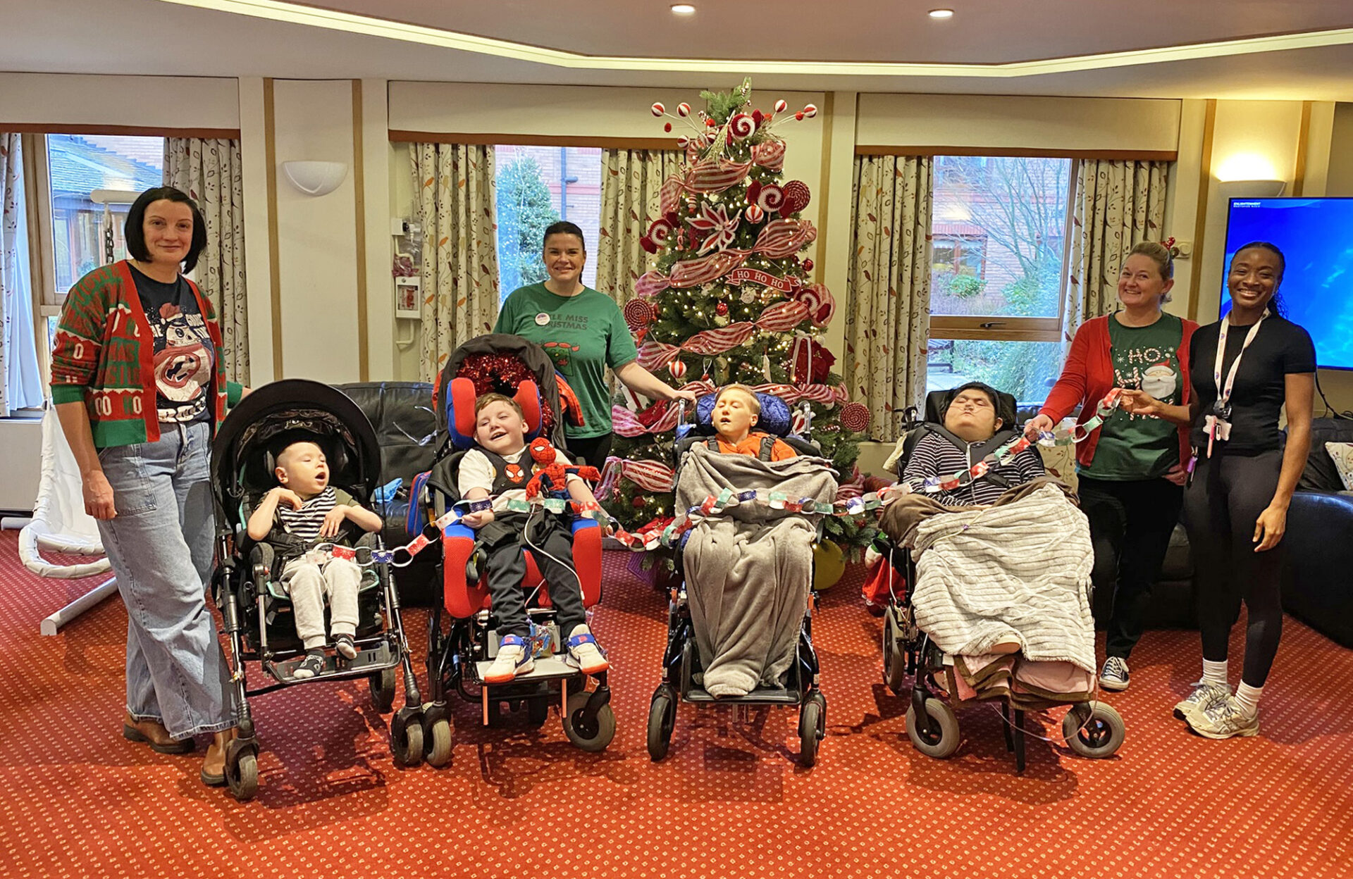Children in wheelchairs and a women wearing Christmas jumpers in front of a Christmas tree holding a garland