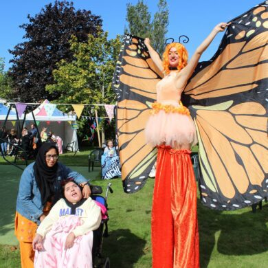 Woman on stilts wearing orange trousers and butterfly wings holding her arms outstretched with a woman stood by a young girl in a wheelchair in a sunny garden