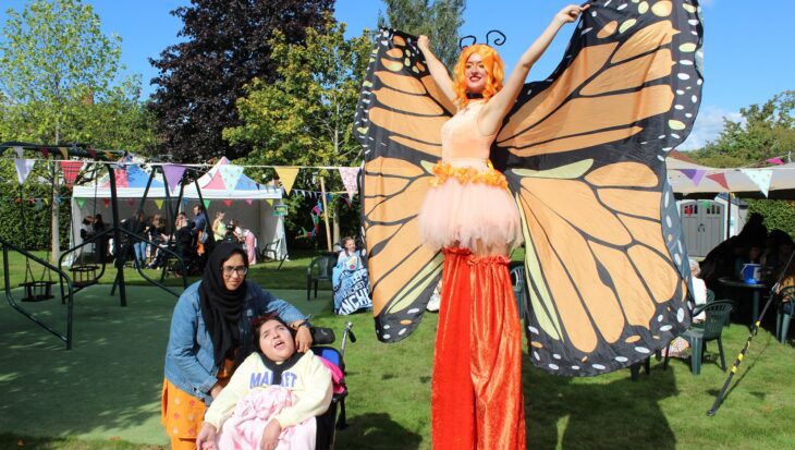Woman on stilts wearing orange trousers and butterfly wings holding her arms outstretched with a woman stood by a young girl in a wheelchair in a sunny garden