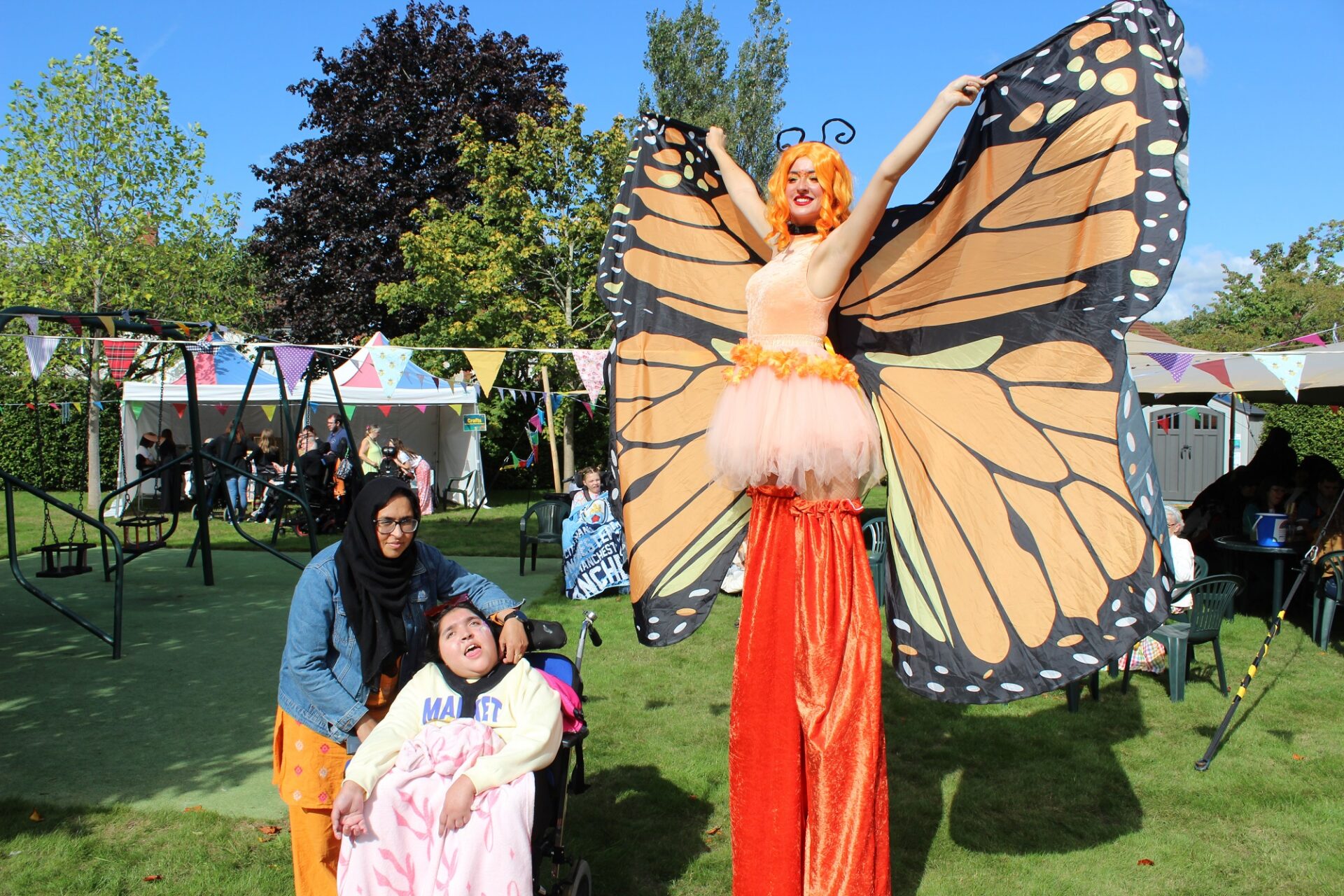 Woman on stilts wearing orange trousers and butterfly wings holding her arms outstretched with a woman stood by a young girl in a wheelchair in a sunny garden