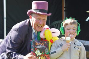 Man wearing a pink top hat wearing a magician outfit and holding balloons next to a young boy wearing green headphones eating an ice cream