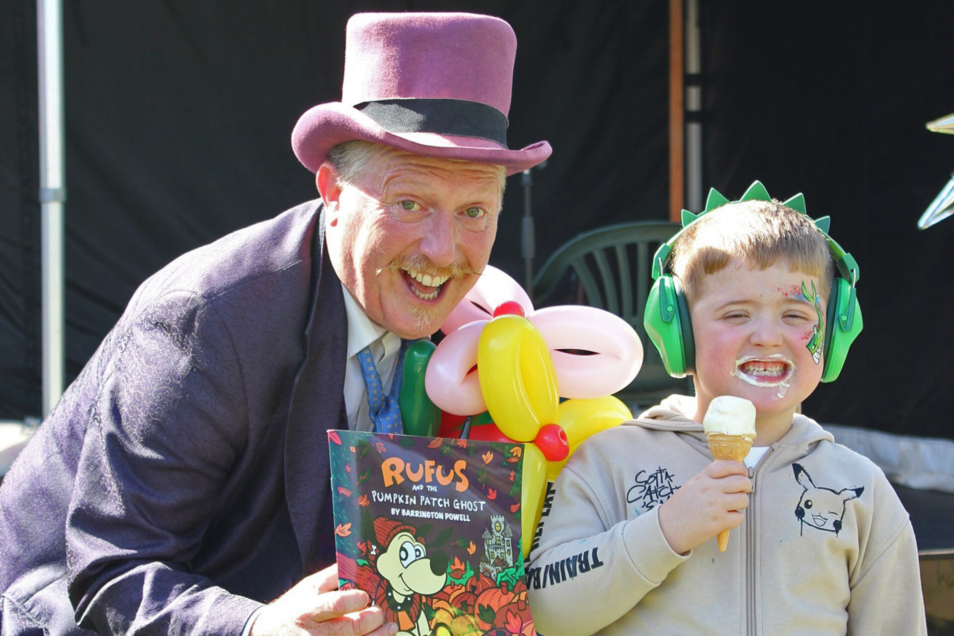 Man wearing a pink top hat wearing a magician outfit and holding balloons next to a young boy wearing green headphones eating an ice cream