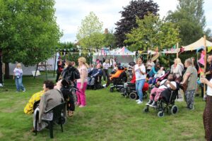 Small crowd of adults stood with young people in wheelchairs in a garden with trees and tents