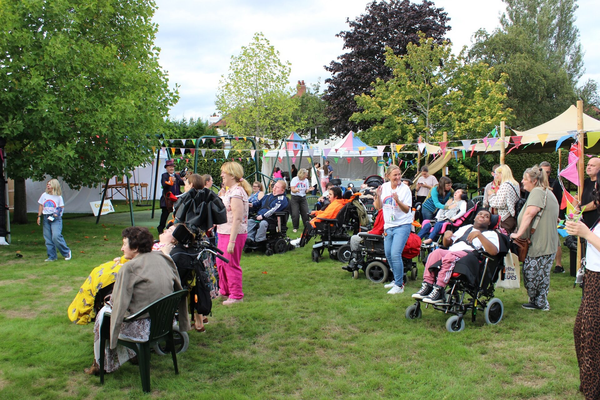 Small crowd of adults stood with young people in wheelchairs in a garden with trees and tents