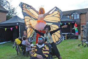Woman on stilts wearing orange trousers and butterfly wings holding her arms outstretched with a woman stood with her arms outstretched stood behind a boy in a wheelchair in a sunny garden