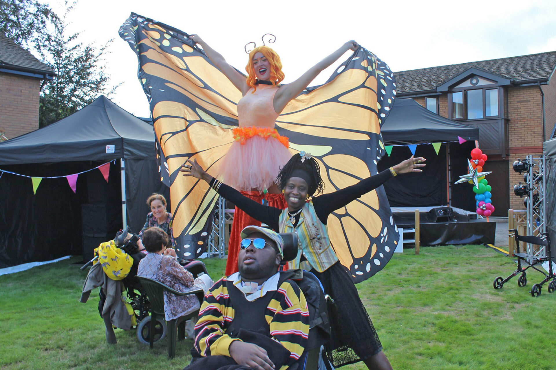 Woman on stilts wearing orange trousers and butterfly wings holding her arms outstretched with a woman stood with her arms outstretched stood behind a boy in a wheelchair in a sunny garden
