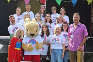 Group of men and women wearing white t-shirts with a rainbow logo and a mouse mascot stood by a stage with colourful bunting