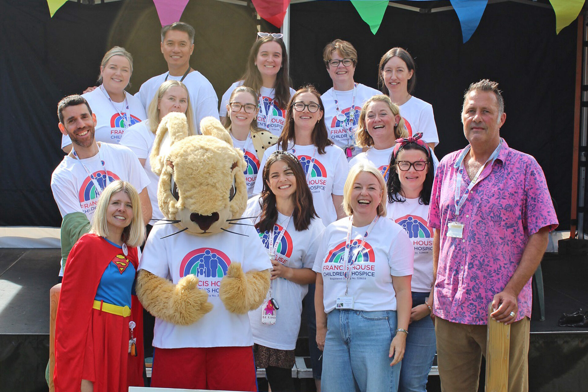 Group of men and women wearing white t-shirts with a rainbow logo and a mouse mascot stood by a stage with colourful bunting