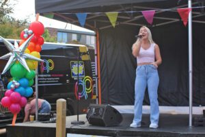 Woman with blonde hair swearing a pink top and pale blue jeans stood singing on a stage with colourful balloons and bunting