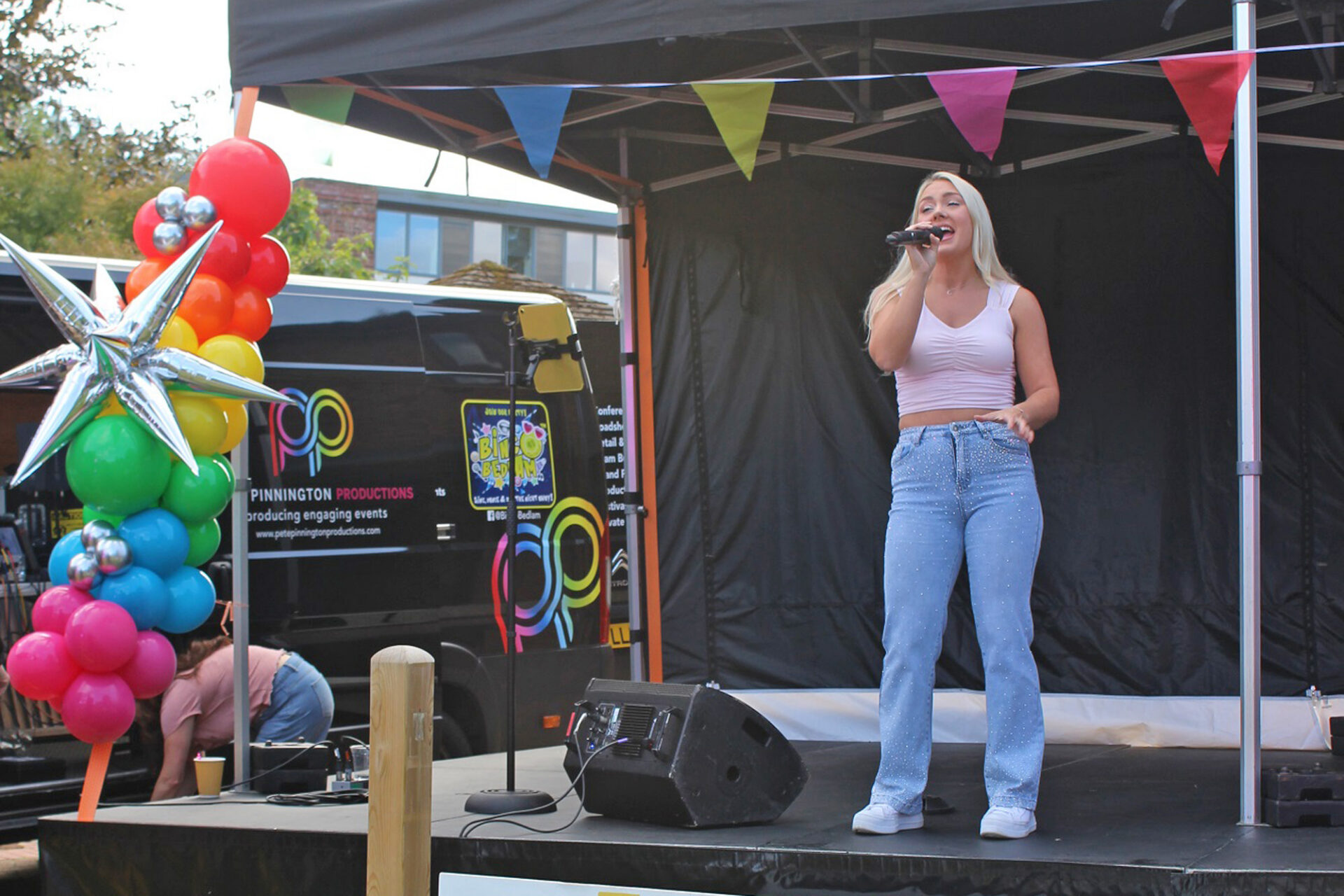 Woman with blonde hair swearing a pink top and pale blue jeans stood singing on a stage with colourful balloons and bunting