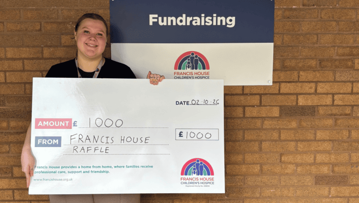 Woman holding a large cheque with the words £1000 pounds stood against a brick wall