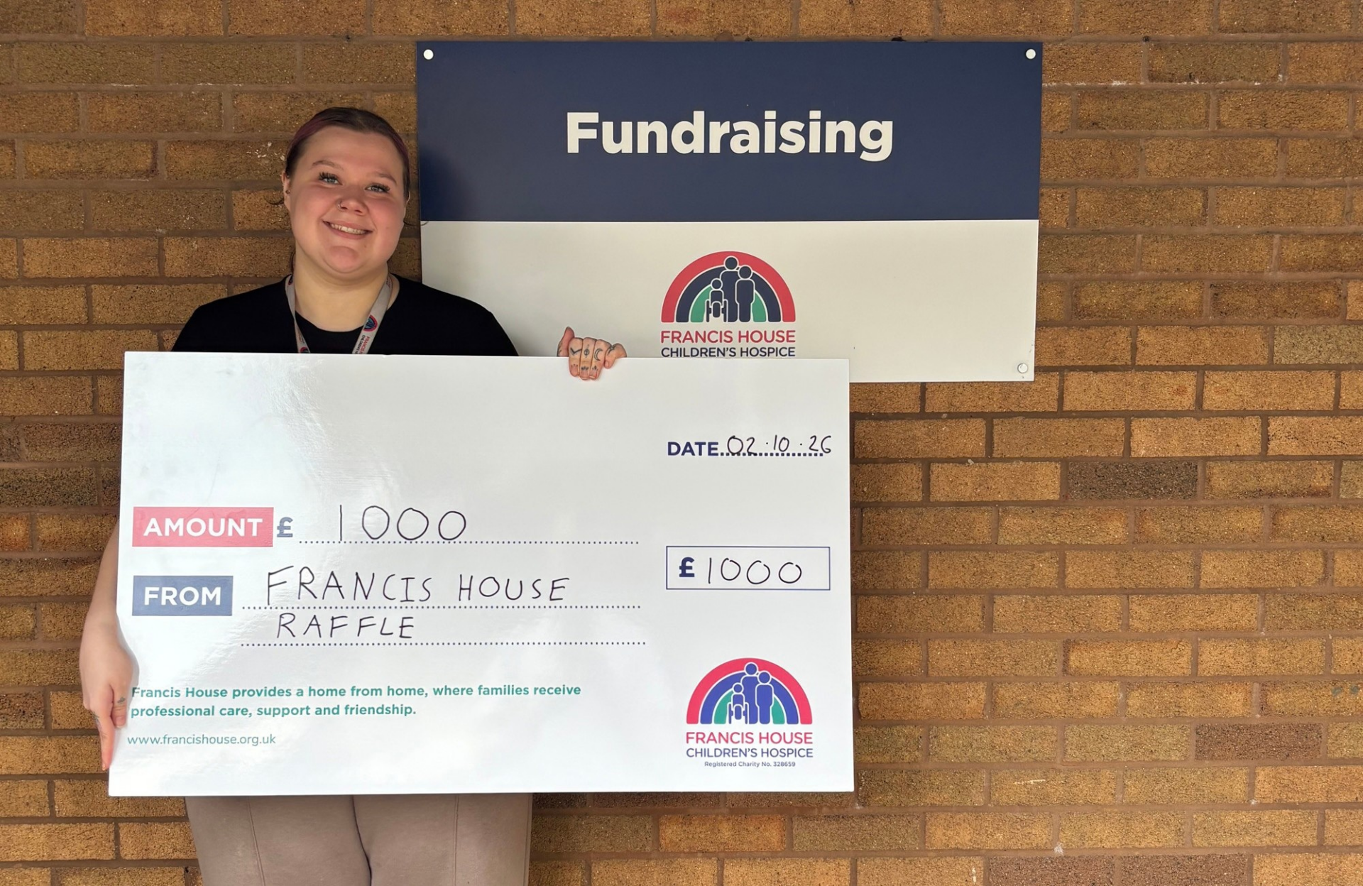Woman holding a large cheque with the words £1000 pounds stood against a brick wall