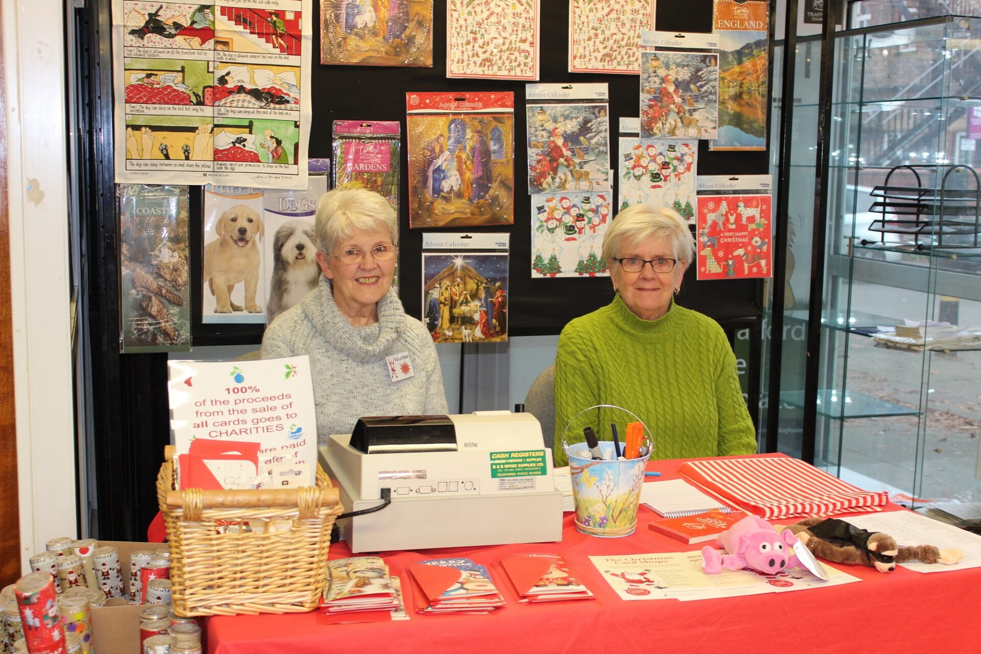 Two women with grey hair wearing jumpers sat in front of a wall of Christmas calendars and behind a table of Christmas gifts and a till.