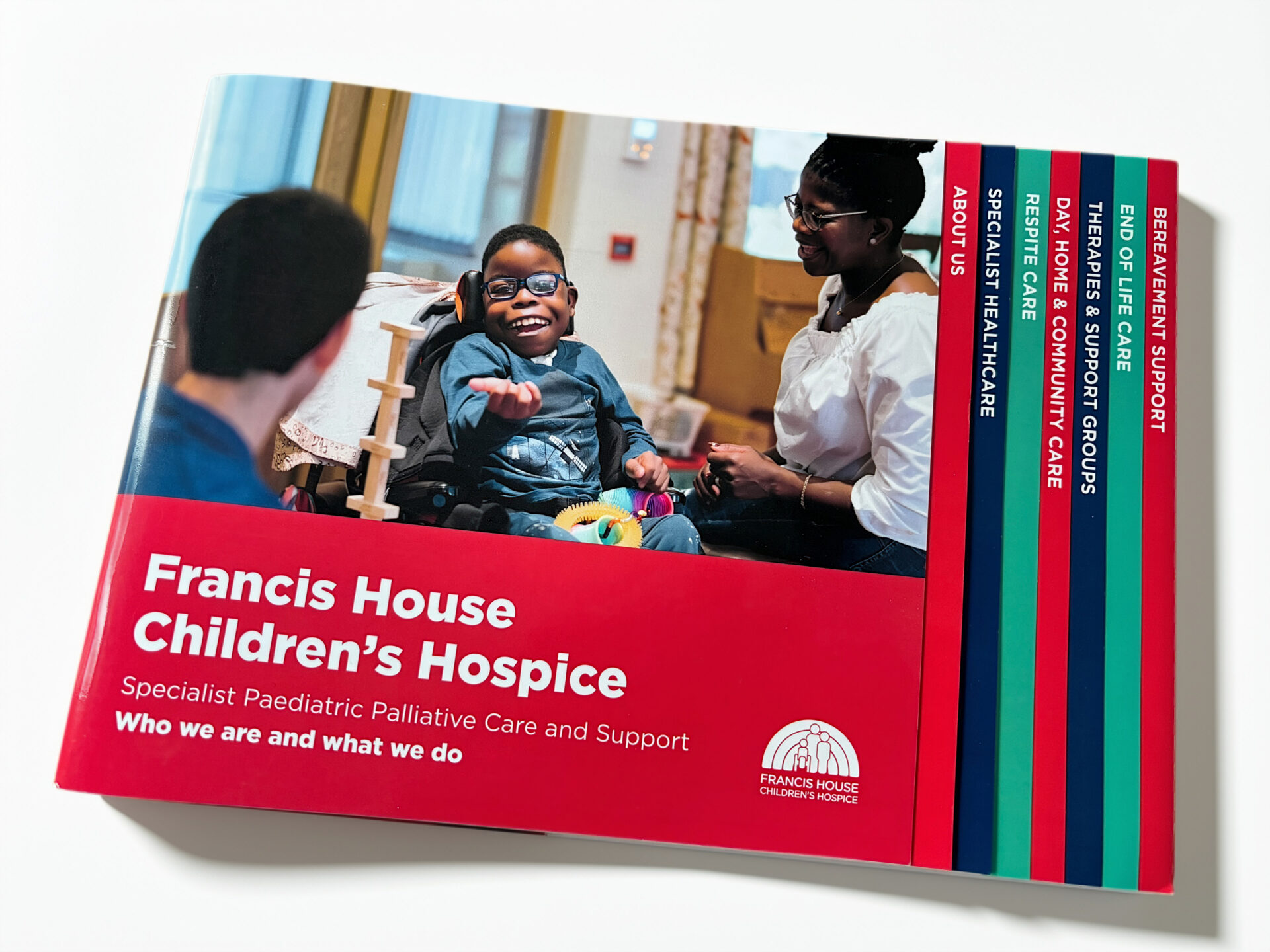 Front cover of a brochure with an image of a child in a wheelchair smiling at the camera with tow adults seated alongside. The title of the brochure appears on a red background with the words Francis House Children's Hospice - who are and what we do.