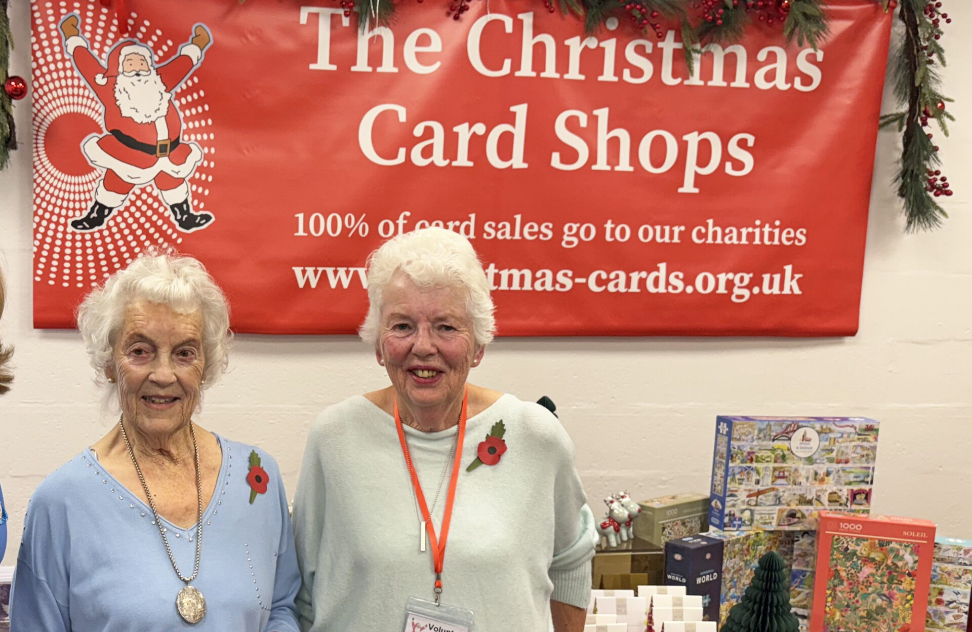 Two women with grey hair wearing jumpers and poppies stood in front of a large banner with the words The Christmas Card Shops and a table of Christmas gifts